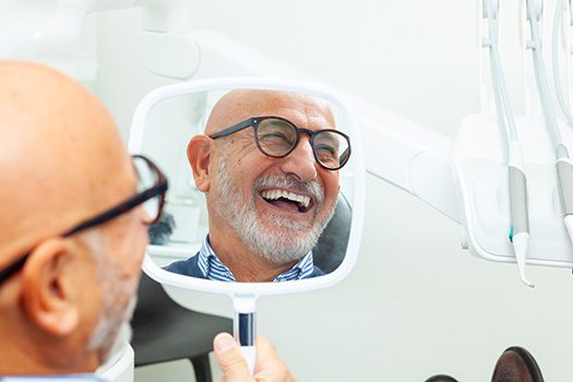 A bald, elderly man smiling while using a hand mirror