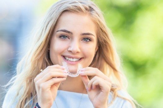 Smiling blonde woman holding Invisalign clear aligner outdoors