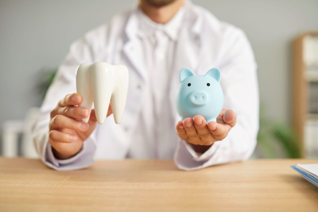 Dentist in white coat holding model tooth and blue piggy bank